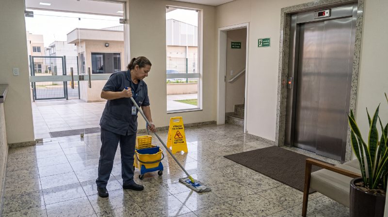Auxiliar de limpeza passando o esfregão no hall ao lado do elevador